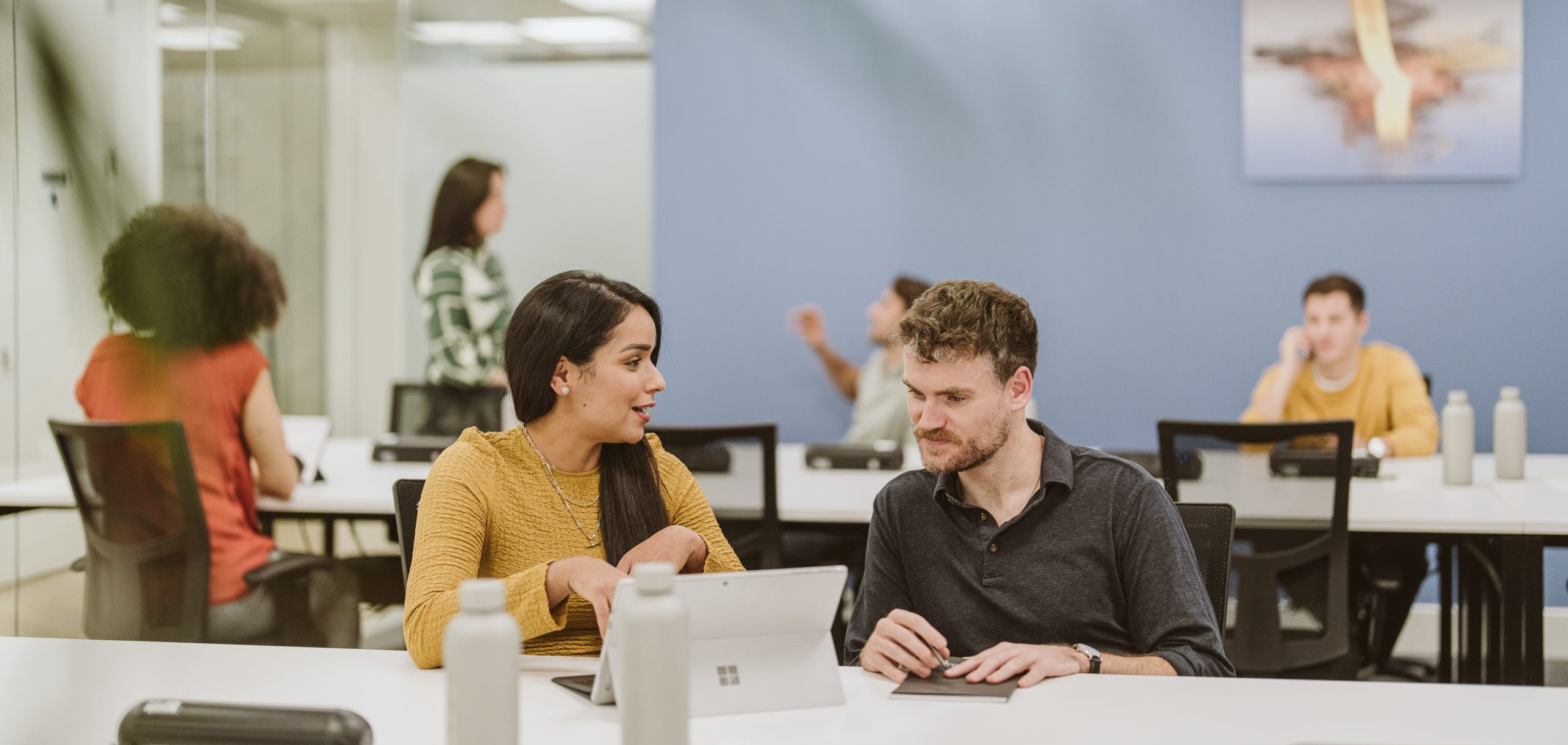 A man and a woman having a discussion at a desk gathered round a laptop with a table of people behind them